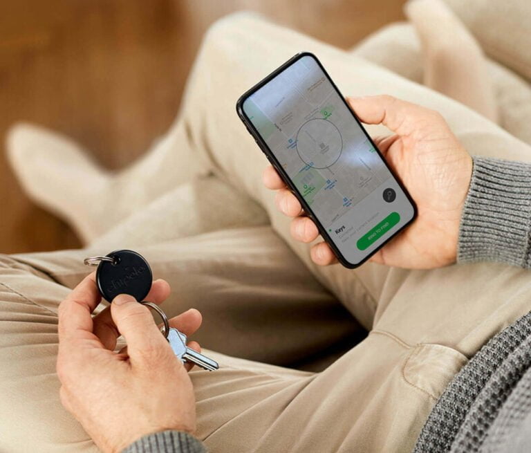 Osoba trzymająca smartfon z otwartą aplikacją do lokalizacji przedmiotów oraz klucze z przyczepionym lokalizatorem Bluetooth w drugiej ręce.