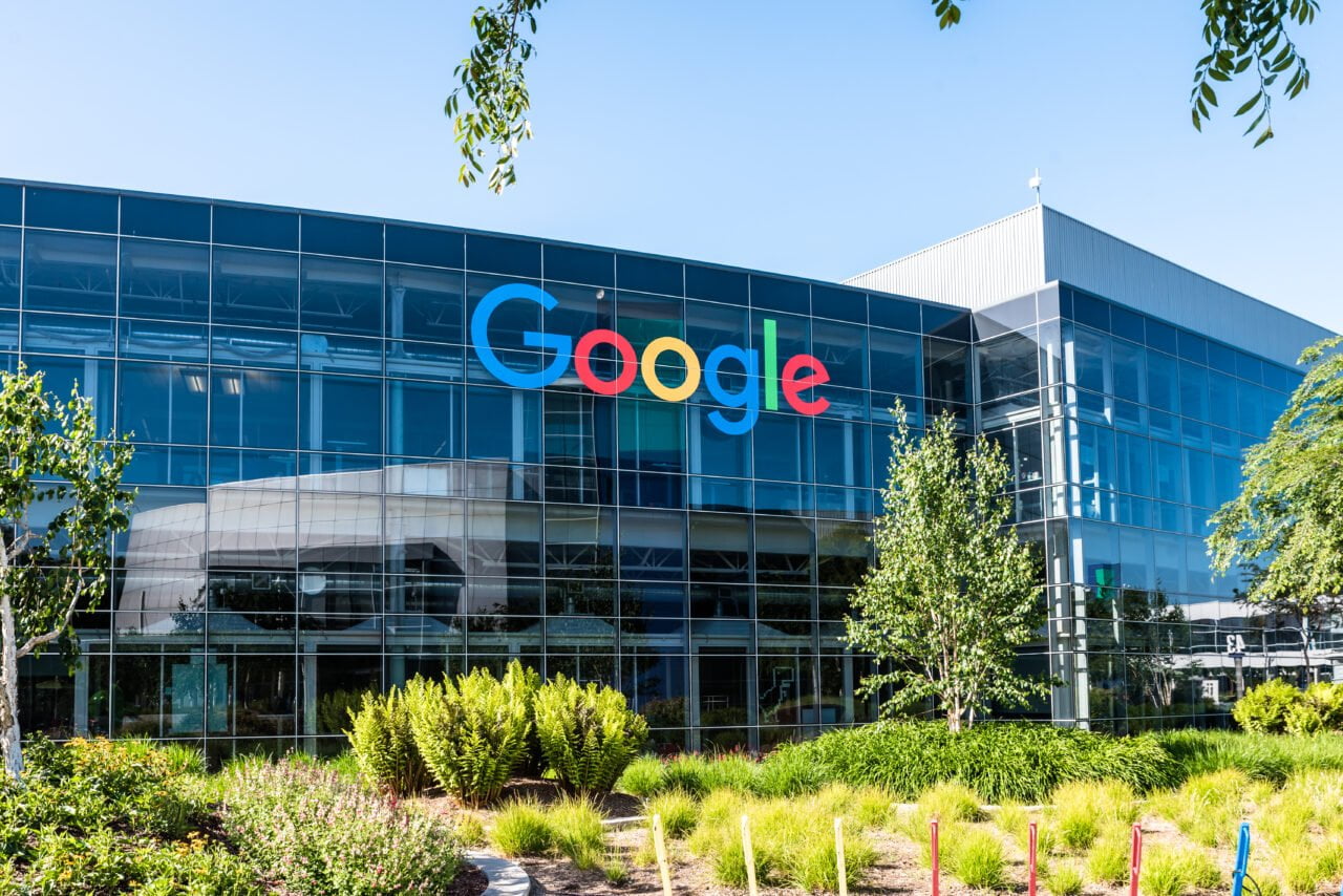 Budynek biurowy Google (Alphabet) z dużym logo firmy na fasadzie, widziany przez zielony teren z drzewami i krzewami w dzień. Chiński pracownik Google został zwolniony z tego miejsca