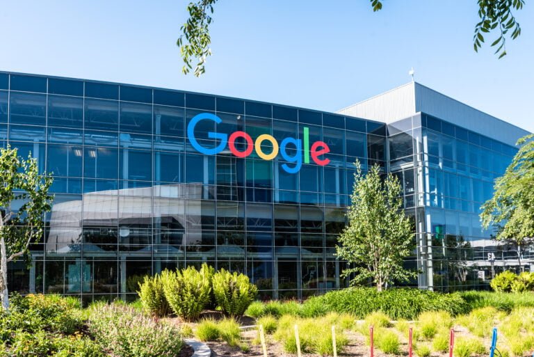 Budynek biurowy Google z dużym logo firmy na fasadzie, widziany przez zielony teren z drzewami i krzewami w dzień. Chiński pracownik Google został zwolniony z tego miejsca