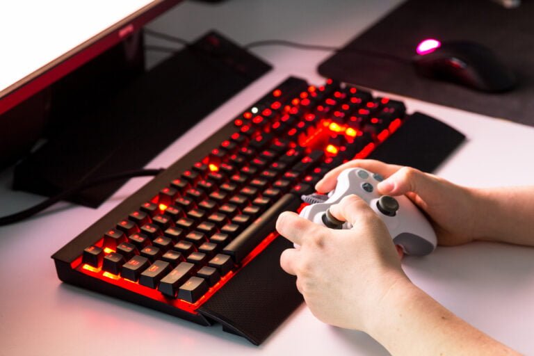 Osoba trzymająca biały kontroler do gier przy czerwono podświetlanej klawiaturze mechanicznej, mysz komputerowa i monitor w tle.