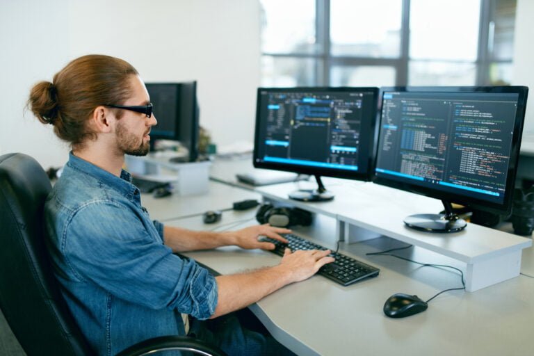 Mężczyzna programista pracujący na dwóch monitorach komputerowych z kodem programistycznym w jasnym biurze. Jakie są wynagrodzenia IT?