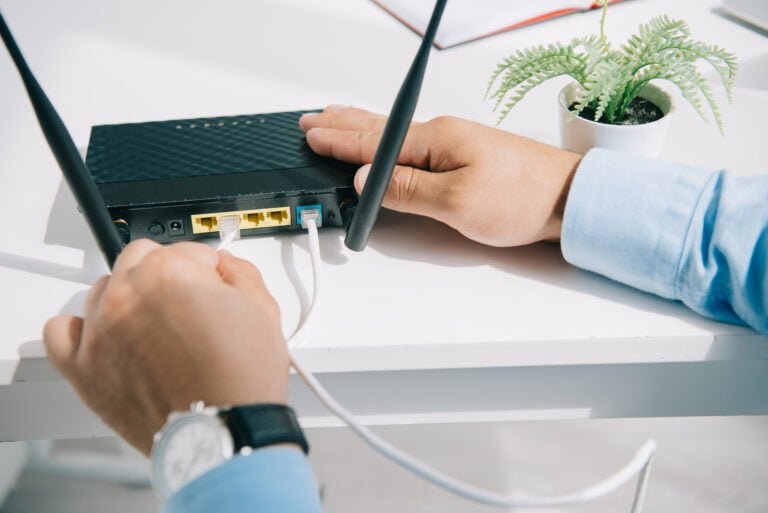 Osoba podłącza kabel sieciowy do routera obok doniczki z zieloną rośliną na biurku.