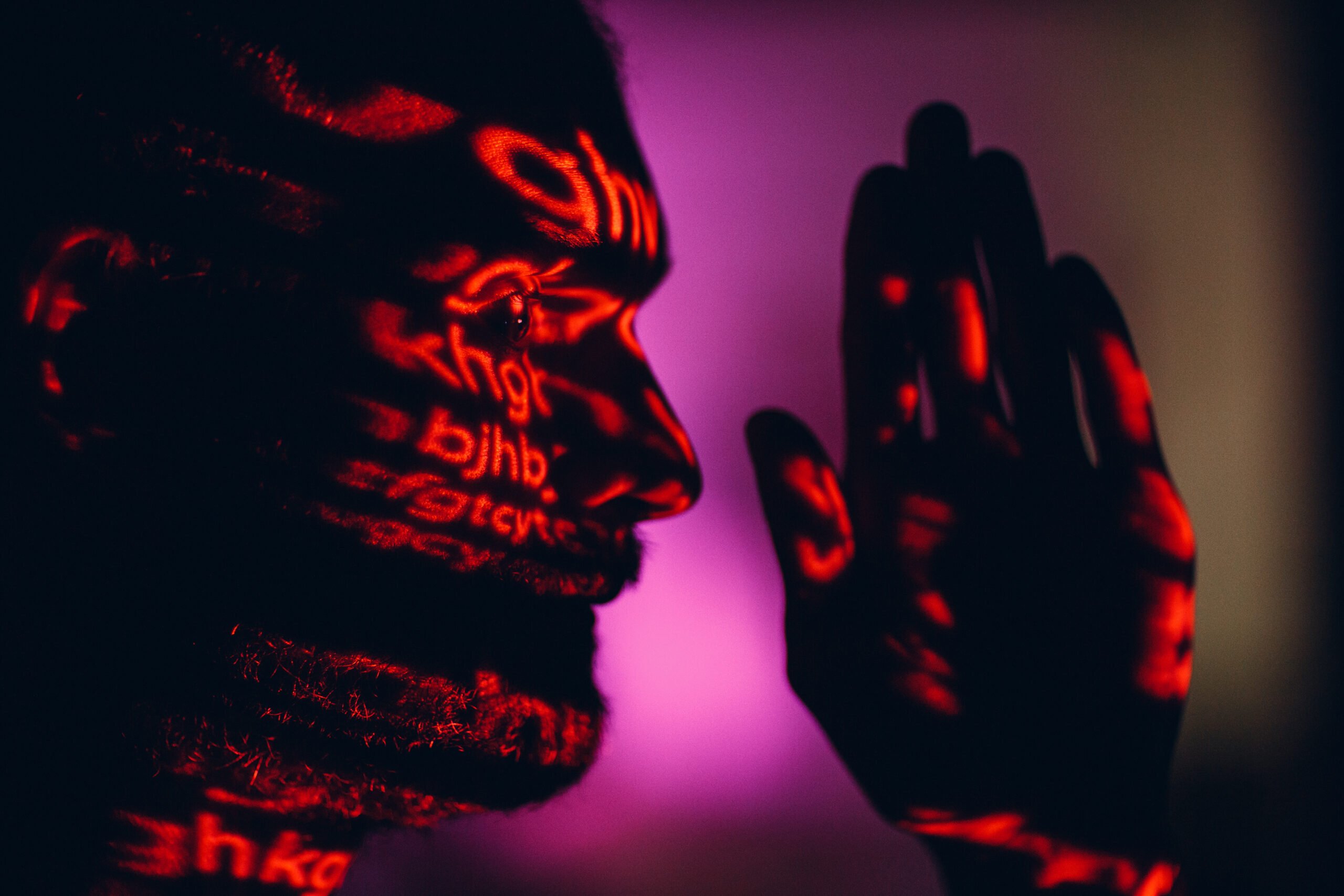 Portret mężczyzny oświetlony czerwonym światłem z napisami rzutowanymi na twarz i dłoń.