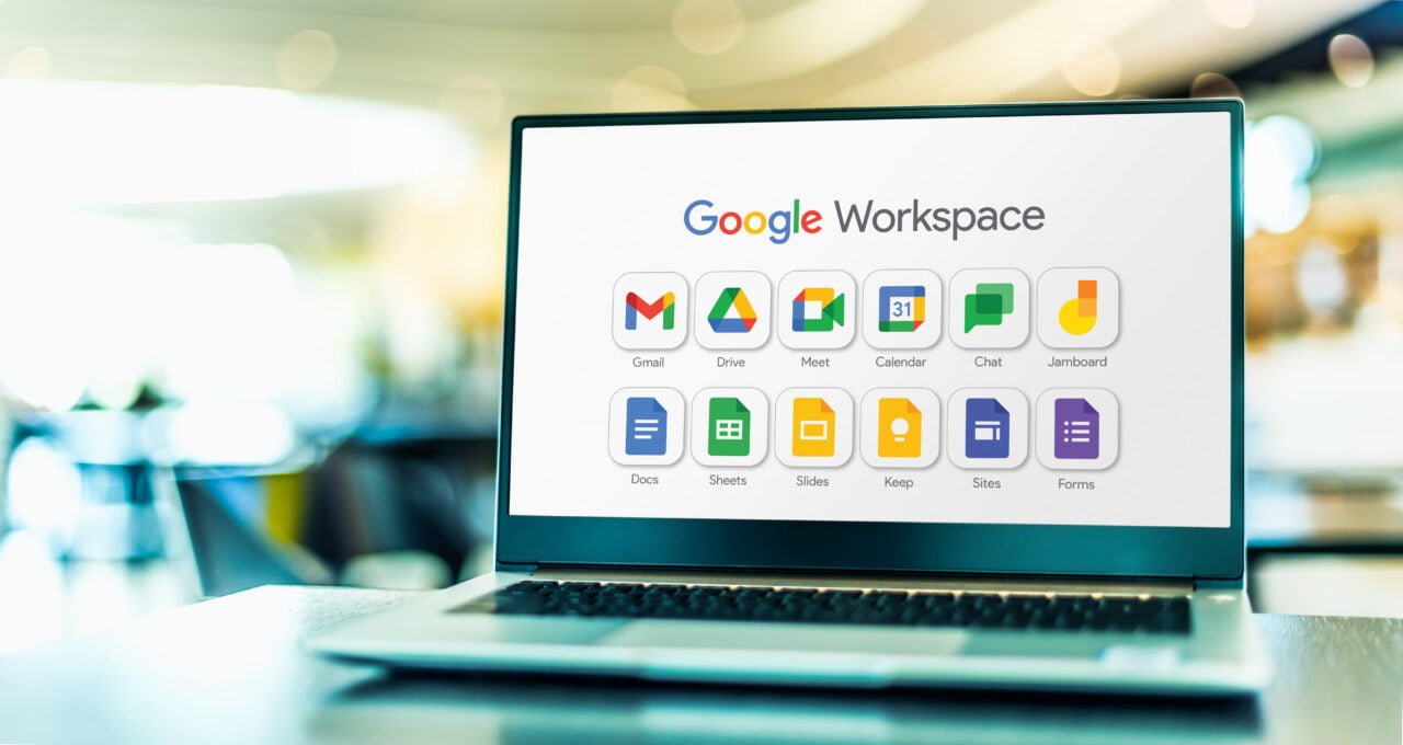 Laptop na stole z ekranem przedstawiającym logo Google Workspace i ikony różnych aplikacji, takich jak Gmail, dysk google, Meet, w tle rozmyte wnętrze.