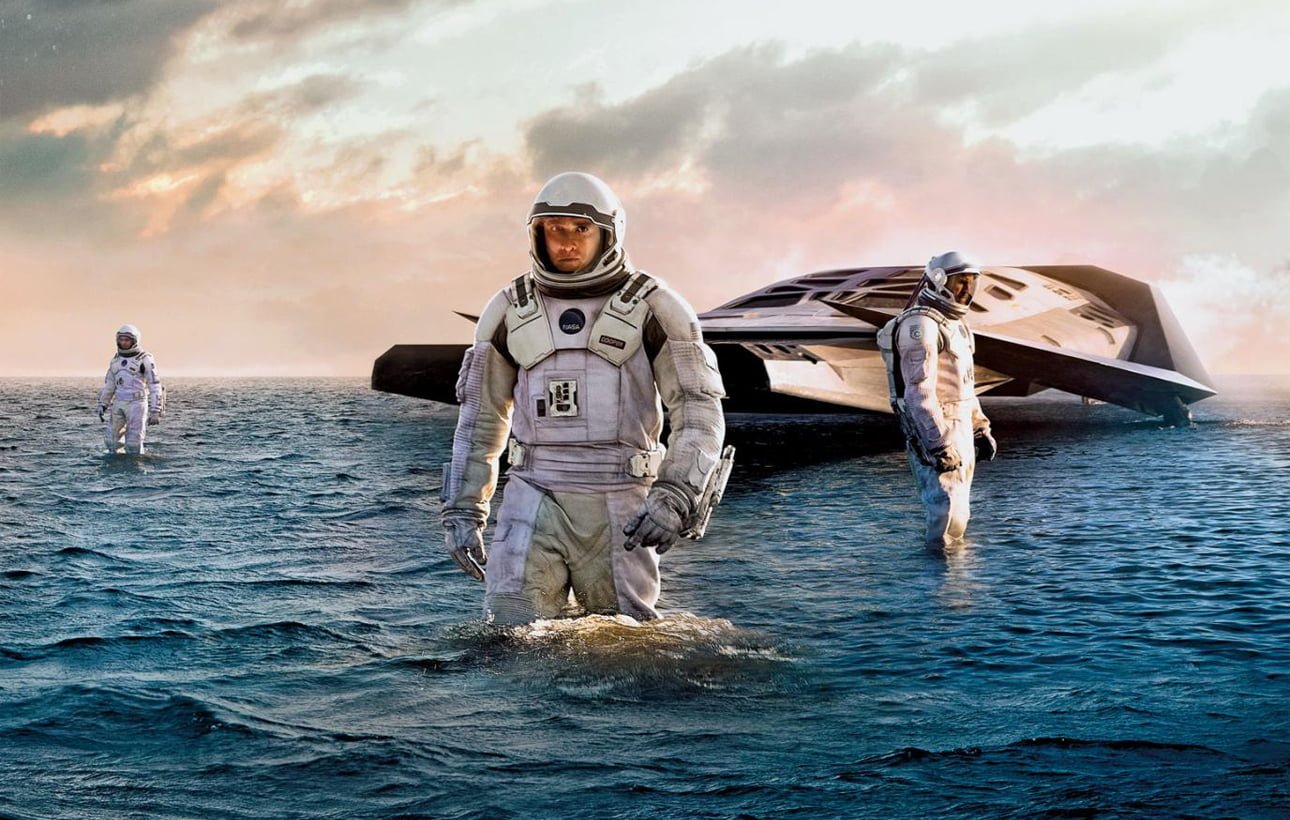 Astronauci w skafandrach kosmicznych na wodzie przed futurystycznym statkiem kosmicznym, tło z zachmurzonym niebem.