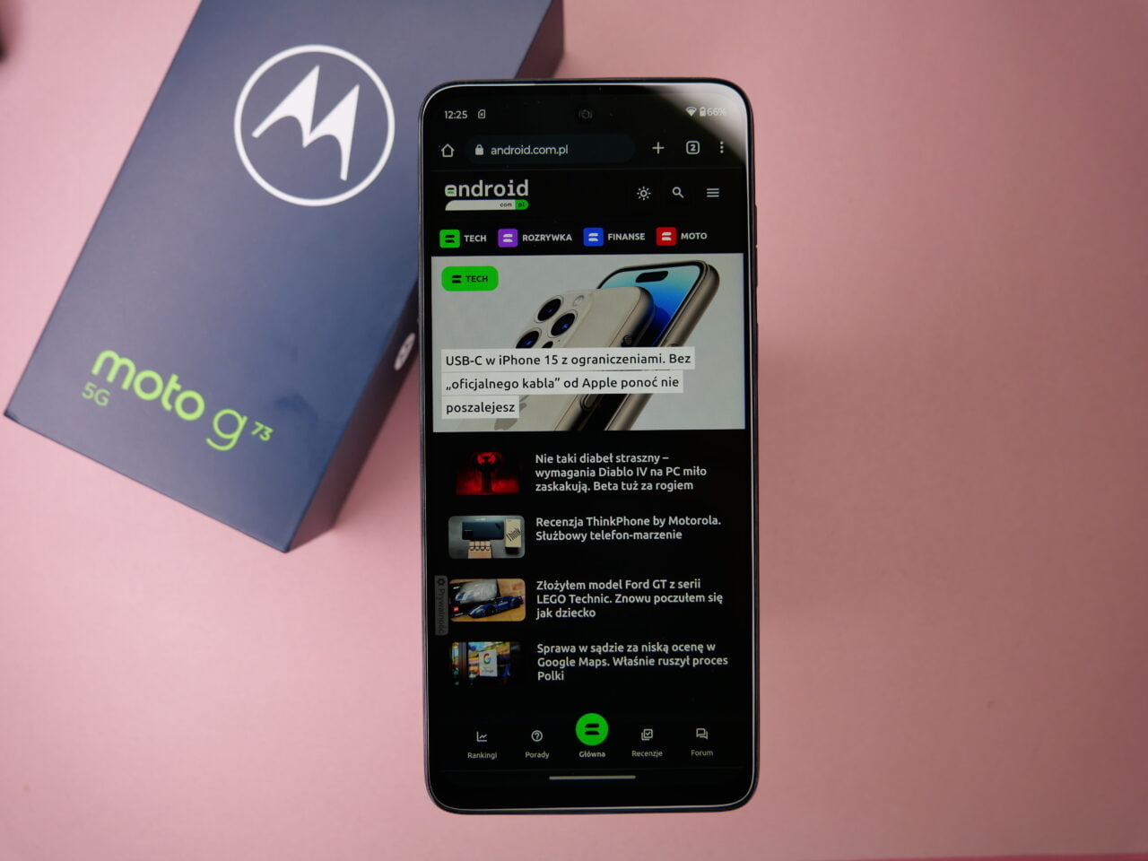 Smartfon Motorola Moto g73 5G leżący na różowym tle obok pudełka z logo Motorola, wyświetlający stronę internetową z artykułami.