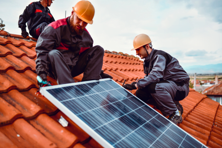 Pracownicy w kaskach instalują panele słoneczne na dachówce na dachu budynku. Domowa fotowoltaika to coraz popularniejszy wybór.