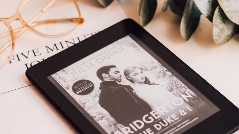 Czytnik e-booków leżący na białym stole, wyświetlający okładkę książki "Bridgerton: The Duke & I" autorstwa Julii Quinn, obok okularów i zielonego kwiatka w doniczce.