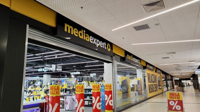 Widok na wejście do sklepu Media Expert w centrum handlowym, z wystawą produktów elektronicznych i reklamami promocyjnymi.