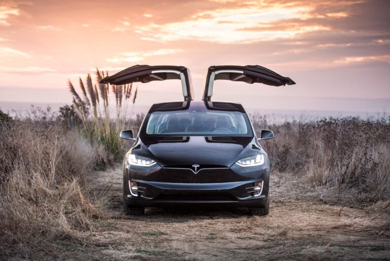 Czarny samochód elektryczny Tesla Model X z otwartymi w górę drzwiami w stylu skrzydeł orła na tle zachodzącego słońca i pola.