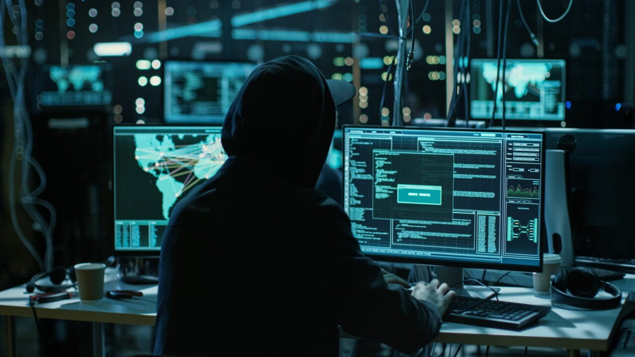 Osoba w kapturze pracująca przy komputerze w ciemnym pomieszczeniu z wieloma monitorami wyświetlającymi kod i dane.