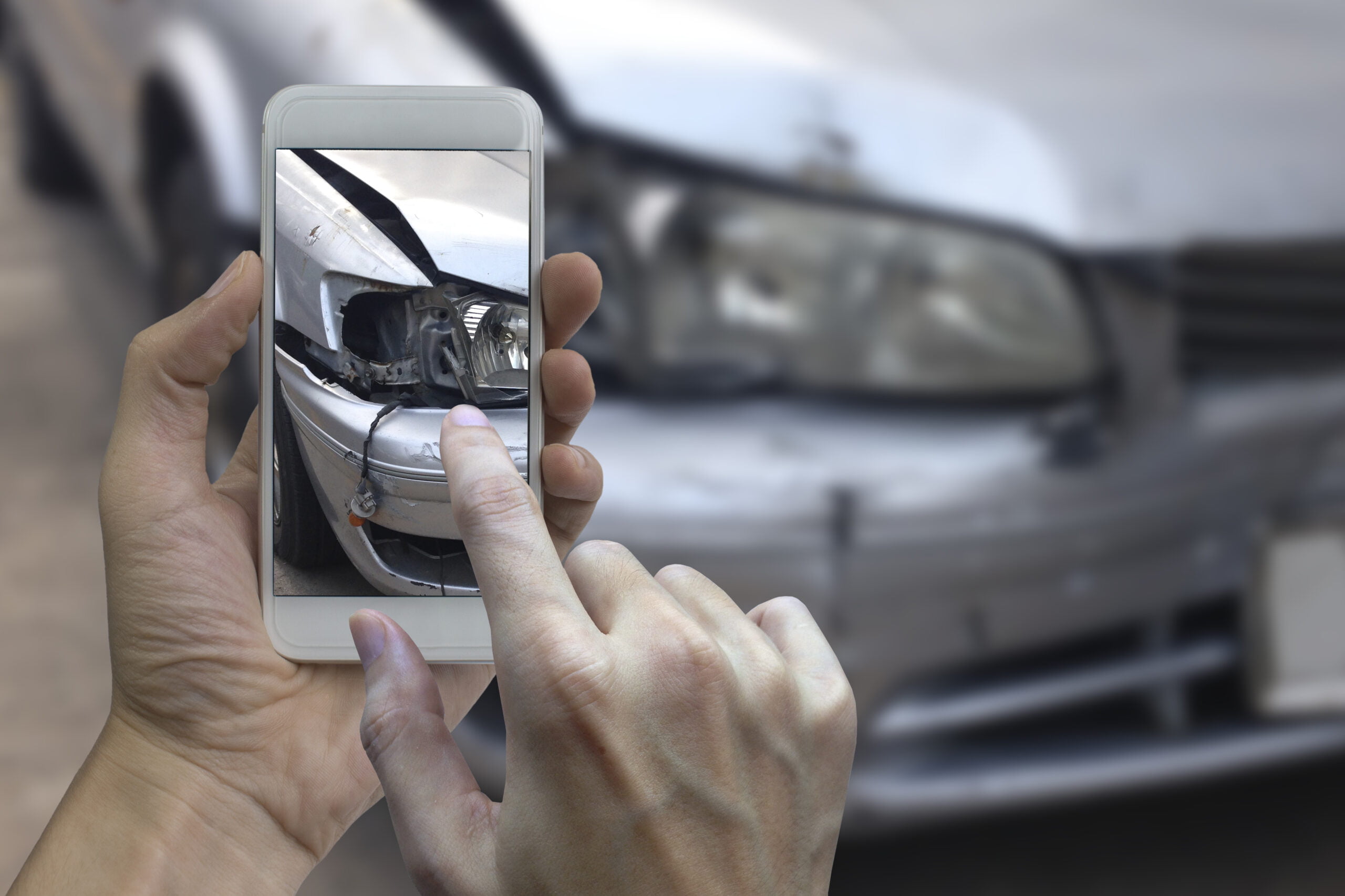 Ręce trzymające smartfon, który robi zdjęcie uszkodzonego przedniego reflektora i zderzaka samochodu.