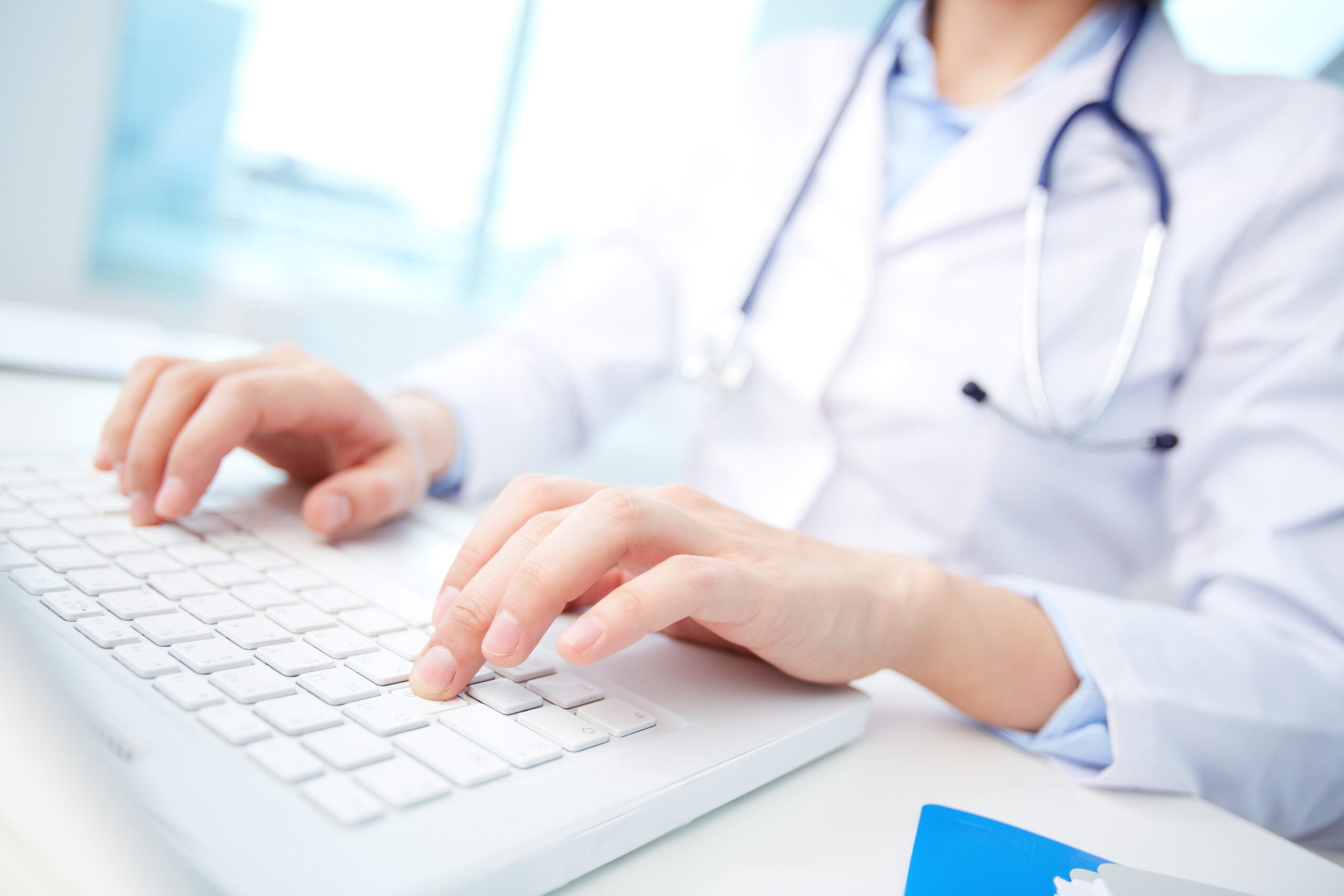 Lekarz w białym fartuchu i ze stetoskopem na szyi pisze na białej klawiaturze laptopa.