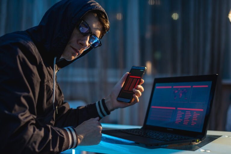 Cyberprzestępcy przy pracy. Osoba w kapturze siedząca przy laptopie i korzystająca z telefonu komórkowego przy bladym świetle nocnego nieba, symbolizująca temat ochrony, jakim jest cyberbezpieczeństwo.