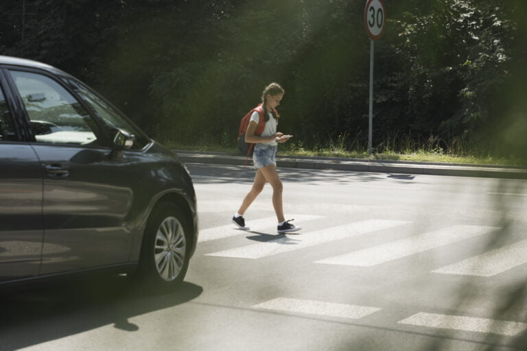 Młoda kobieta z plecakiem przechodzi przez jezdnię z telefonem komórkowym w dłoni, obok jest samochód, a w tle znak ograniczenia prędkości do 30 km/h.