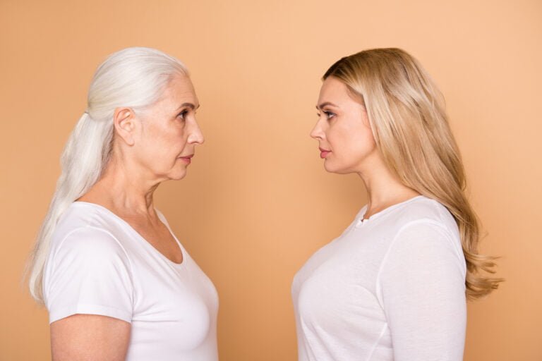 Starsza kobieta o srebrzystych włosach i młodsza kobieta o blond włosach w białych koszulkach stoją naprzeciwko siebie na pomarańczowym tle, patrząc na siebie profilami.