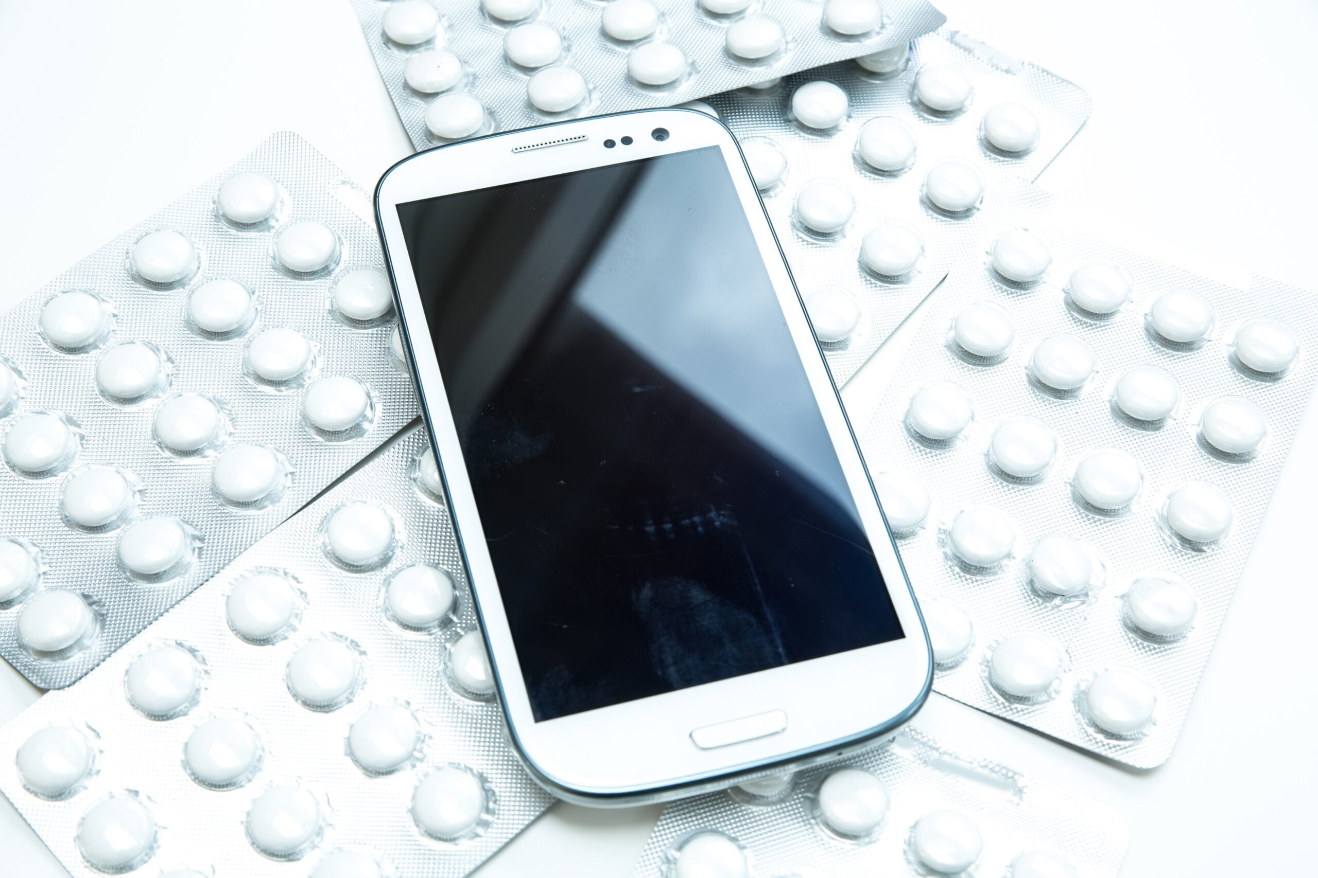 Biały smartfon położony na tle blistrów z tabletkami na białym stole. Aplikacje zdrowotne na twoim smartfonie.