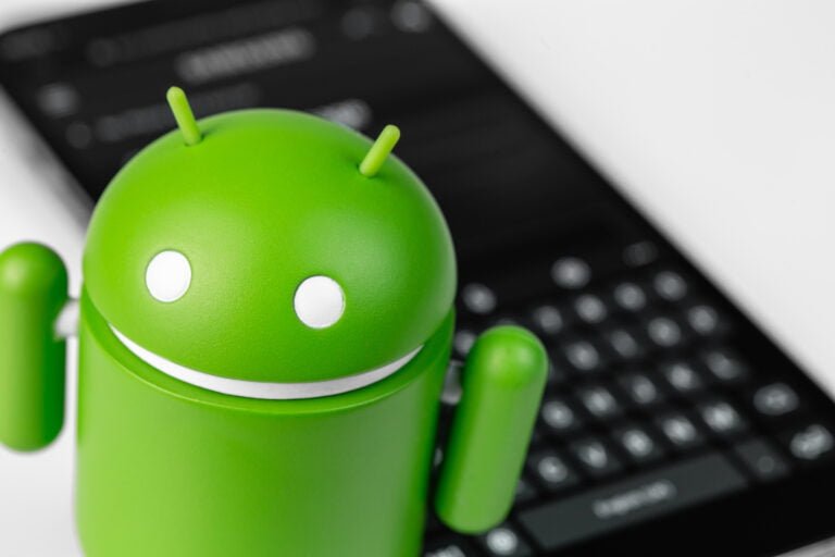 Figurka maskotki Androida w kolorze zielonym na pierwszym planie, w tle częściowo widoczny czarny smartfon z klawiaturą QWERTY.