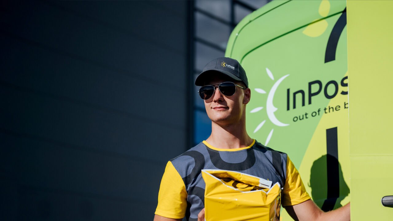kurier InPost w okularach przeciwsłonecznych i czapce z daszkiem, trzymający paczkę, w tle samochód dostawczy w zielonym kolorze z logiem InPost