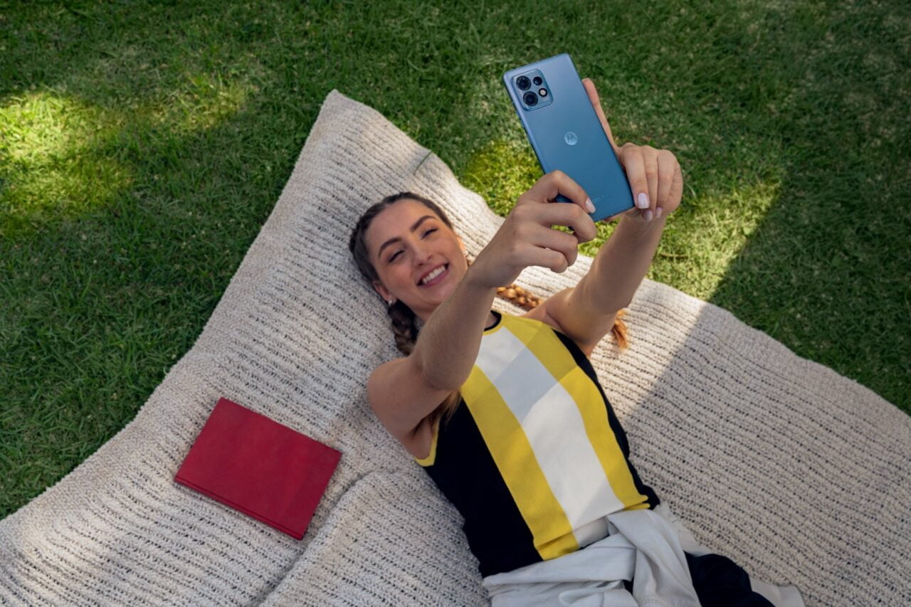 smartfon Motorola edge 40 Pro, Młoda kobieta leżąca na plecach na kocu robi sobie selfie smartfonem na zewnątrz. Obok niej czerwona okładka książki.