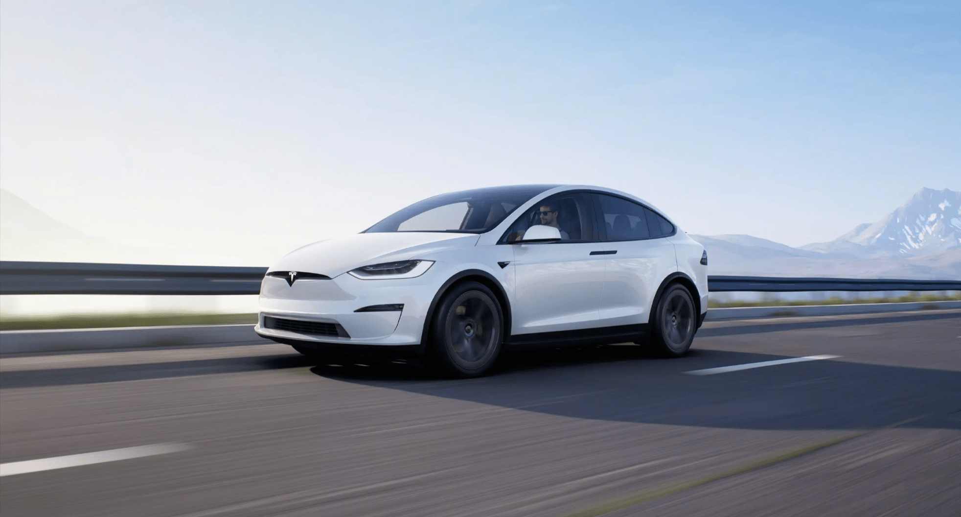 Biały samochód elektryczny Tesla Model X jedzie po autostradzie z rozmytym tłem przedstawiającym góry i niebo.