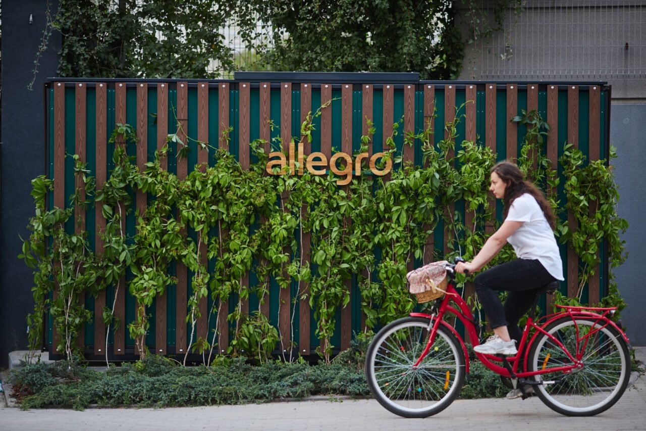 automat paczkowy allegro porośnięty roślinnością, przed nim kobieta jedzie na czerwonym rowerze