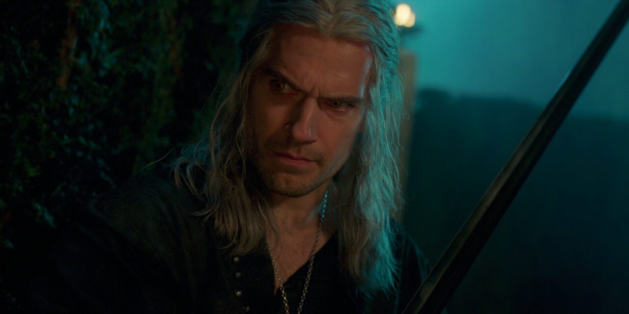 Kadr z serialu Wiedźmin. Geralt z Rivii o długich, białych włosach i intensywnym spojrzeniu, trzymający miecz, na tle mrocznego lasu.