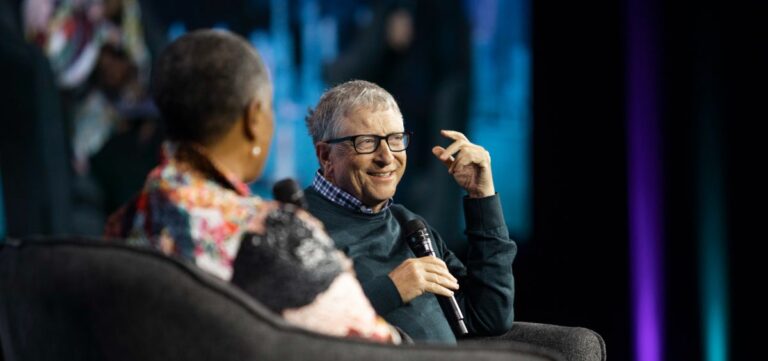 Bill Gates i kobieta siedzą na scenie podczas wydarzenia publicznego; mężczyzna uśmiecha się i gestykuluje, trzymając mikrofon.