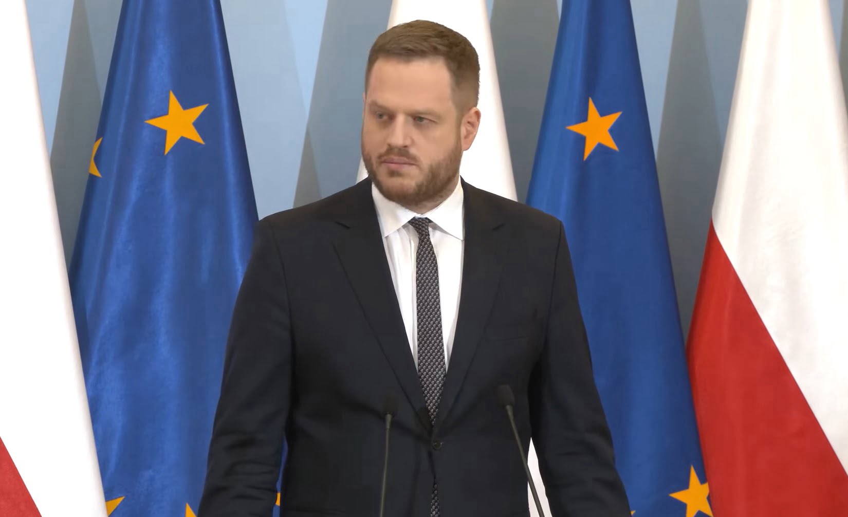 Janusz Cieszyński w garniturze stoi przed mikrofonami z flagami Unii Europejskiej i Polski w tle.