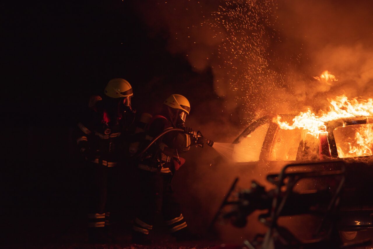 Strażacy gaszący pożar samochodu w nocy, iskry unoszą się nad płomieniami.