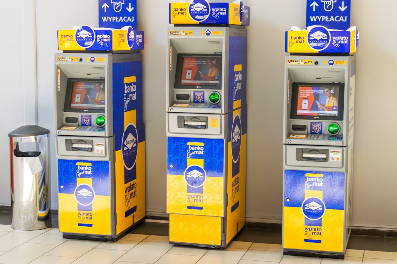 Trzy bankomaty Eurocash z niebiesko-żółtym brandingiem w pomieszczeniu, obok kosz na śmieci.