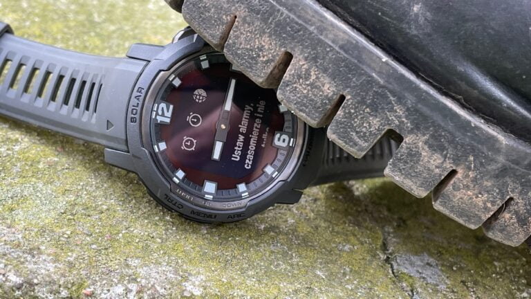Zegarek na ładowanie słoneczne z cyfrowym wyświetlaczem wskazującym różne funkcje po polsku, oparty o ciemne opony na tle kamiennego podłoża.