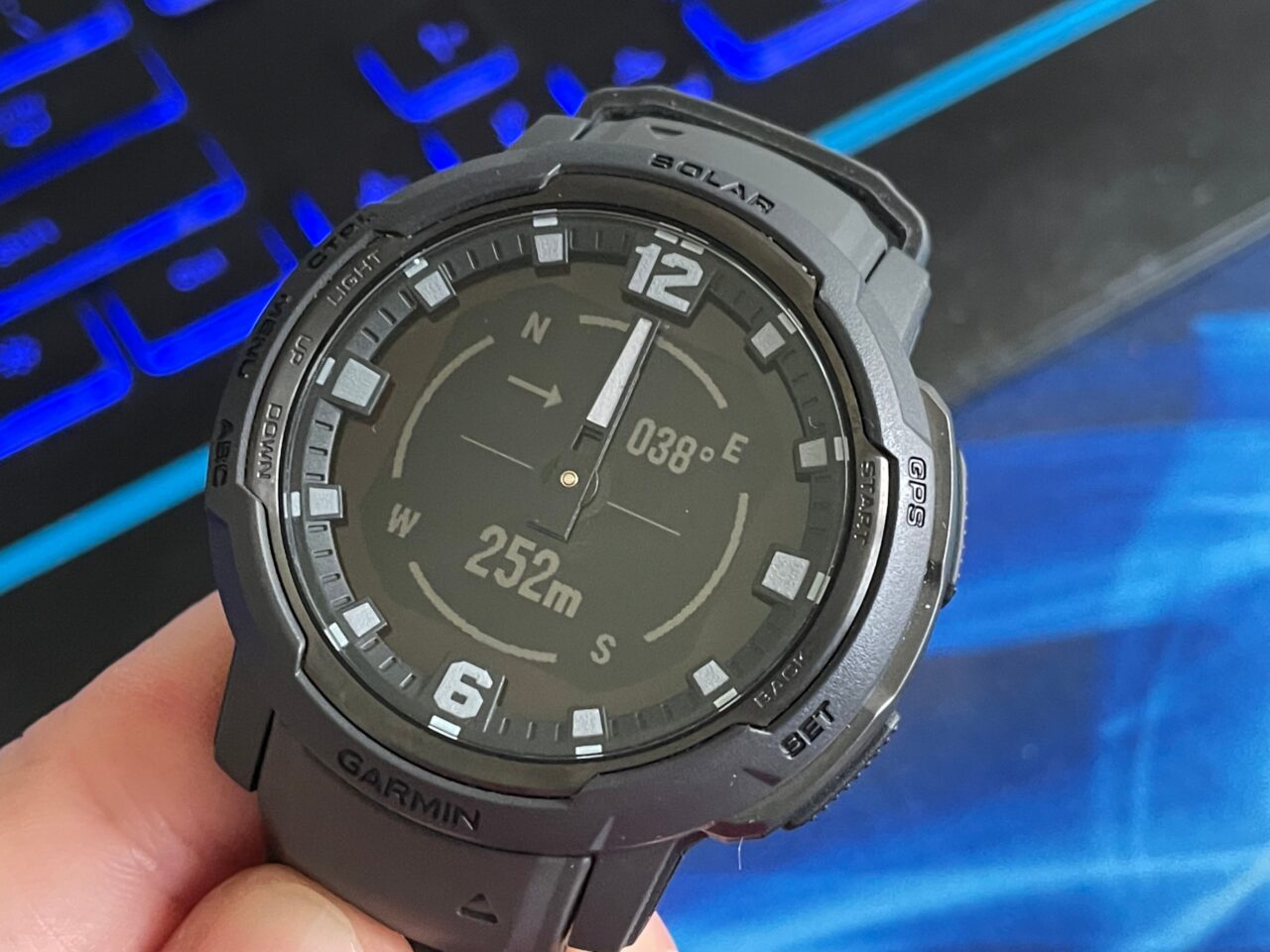 Czarny cyfrowy zegarek sportowy marki Garmin na tle podświetlanej klawiatury komputerowej, wyświetlający kierunek oraz wysokość nad poziomem morza.