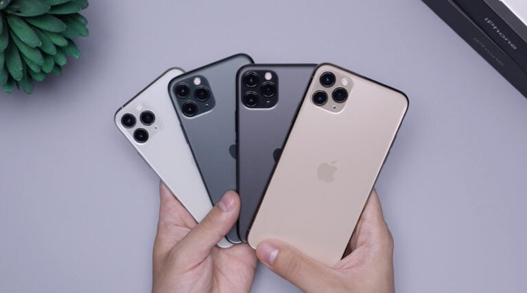 Cztery smartfony iPhone ułożone jedno na drugim o różnych kolorach obudowy, trzymane w dłoni nad szarym stołem, w lewym górnym rogu zielona roślina doniczkowa.
