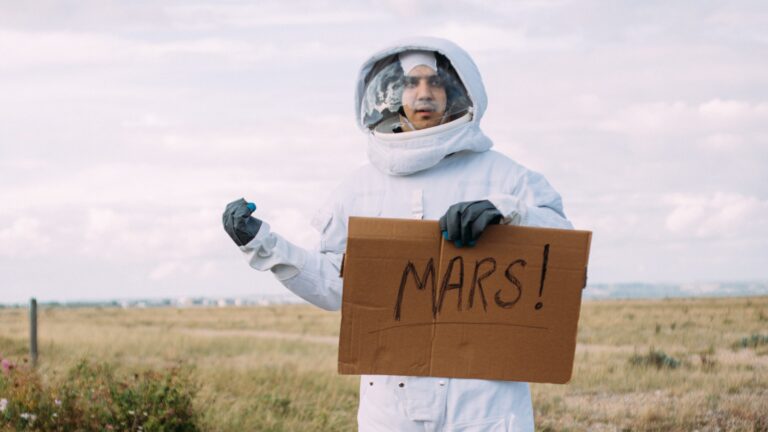 Osoba w skafandrze astronauty trzyma karton z napisem "MARS!" na tle rozległego pola.