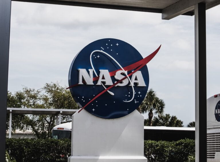 Zdjęcie niebieskiego logo NASA z charakterystycznym czerwonym wektorem na tle częściowo zachmurzonego nieba.