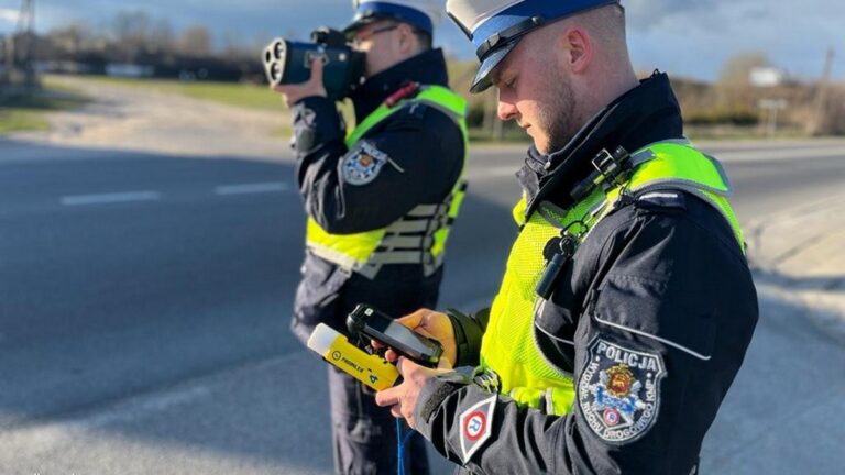 Dwóch policjantów w odblaskowych kamizelkach przeprowadza kontrolę prędkości przy użyciu radaru drogowego na polskiej drodze.