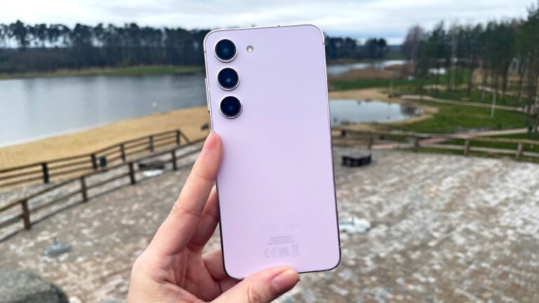 Ręka trzymająca różowy smartfon Samsung Galaxy S23 z potrójnym aparatem w tle krajobraz z nieostrym jeziorem i drzewami.