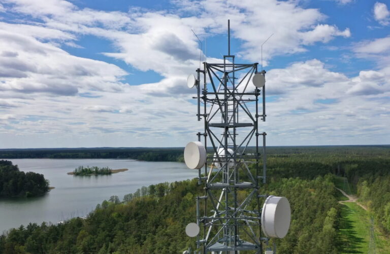 Widok na stalową wieżę telekomunikacyjną z antenami i parabolami na tle niebieskiego nieba, chmur oraz krajobrazu z jeziorem i lasem.