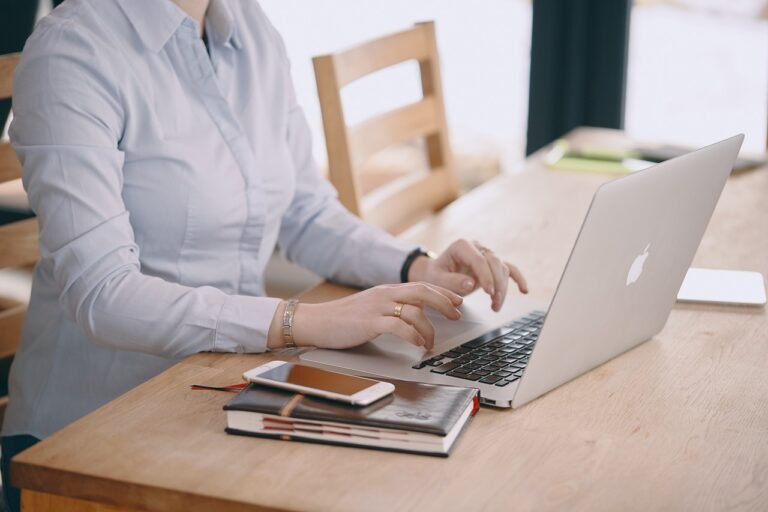 Kobieta pracująca na laptopie marki Apple, siedząca przy drewnianym stole, obok zeszyt i smartfon.