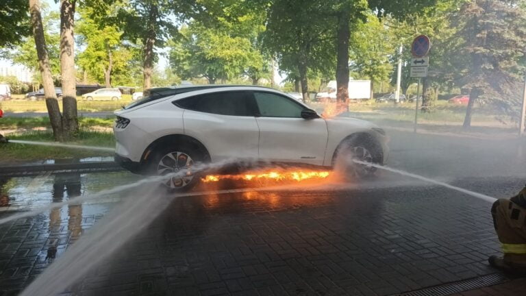Biały samochód elektryczny Mustang Mach-E stoi płonący na parkingu podczas gdy strażak gasi pożar, tryskając wodą z węża strażackiego na płonący spód pojazdu.