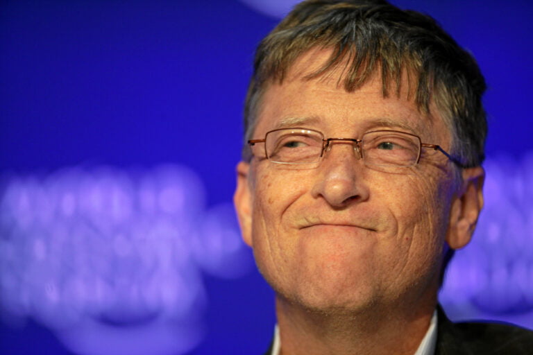 Bill Gates w średnim wieku z okularami, uśmiechający się lekko przed rozmytym niebieskim tłem.