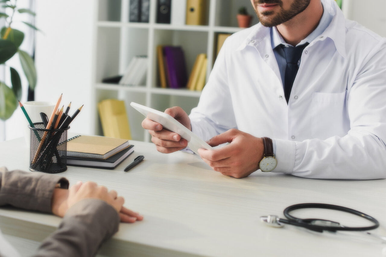 Zmiany w e-receptach. Lekarz w białym fartuchu trzymający tablet w przychodni, z biurkiem z przyborami do pisania i notesami oraz pacjentem siedzącym po przeciwnej stronie. Choroba Parkinsona i konsultacja