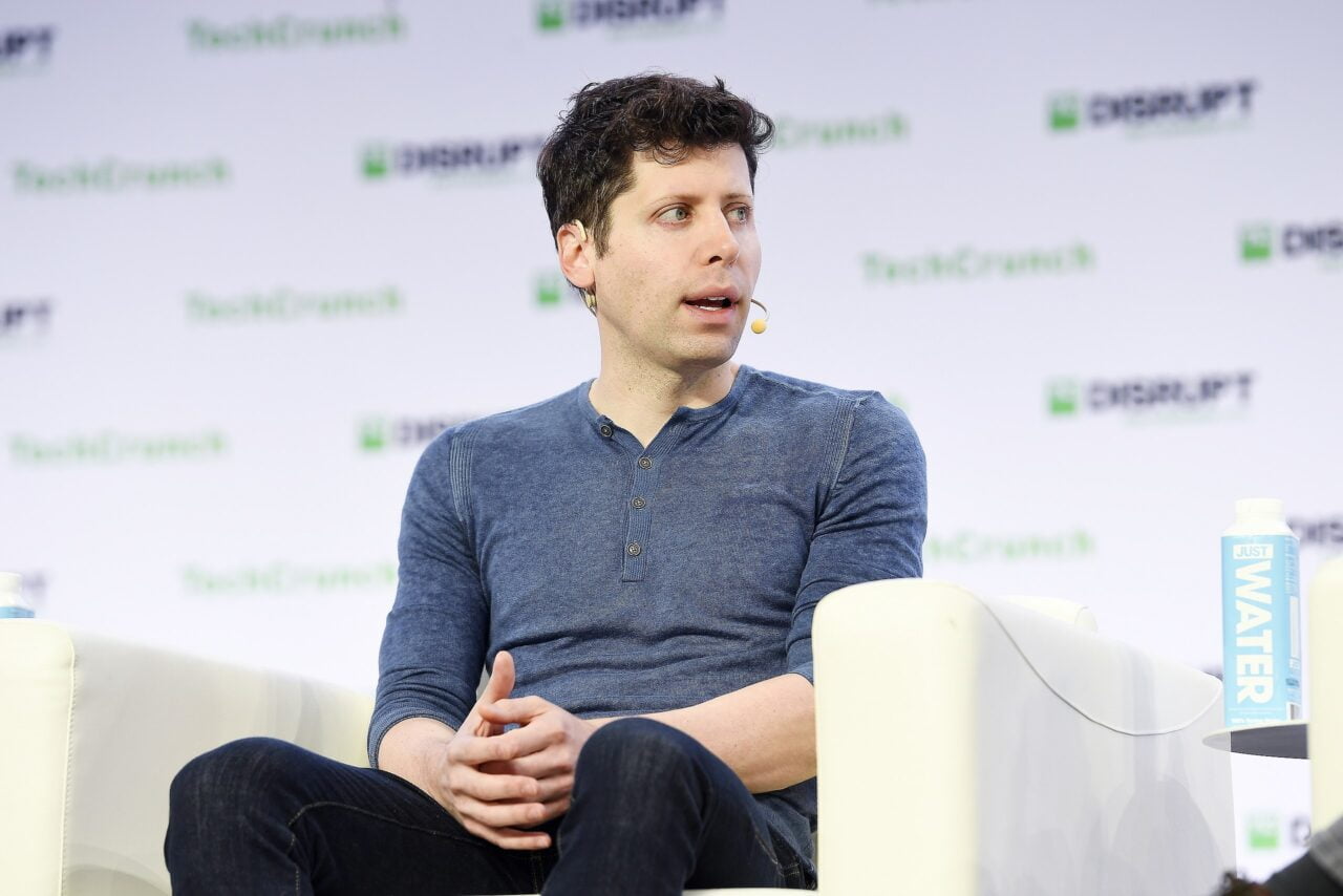 Mężczyzna w niebieskiej koszulce z mikrofonem do ust, siedzący na białym krześle, w tle logo TechCrunch.