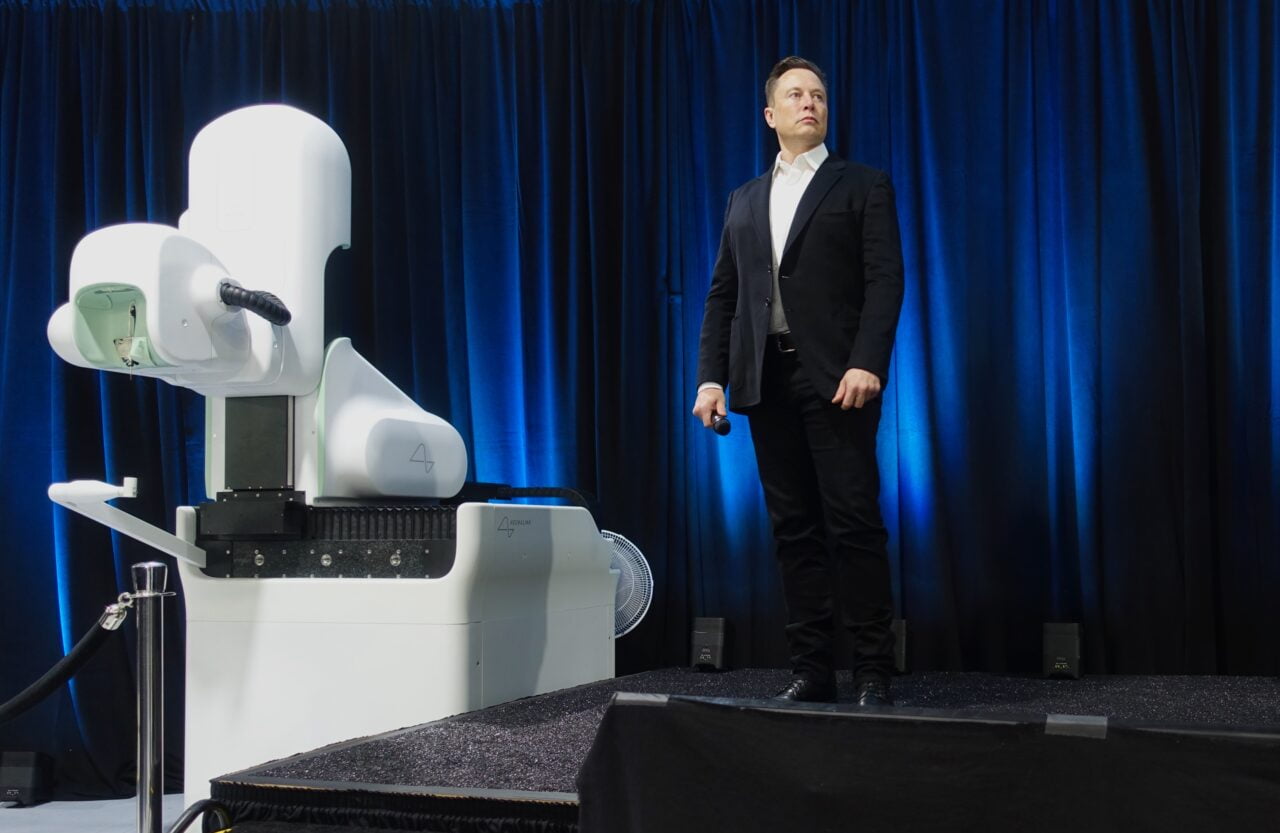 Pierwszy Pacjent Neuralink. Elon Musk, ojciec projektu Nerualink. Mężczyzna w garniturze stoi na scenie obok zaawansowanego technologicznie urządzenia z robotycznym ramieniem.