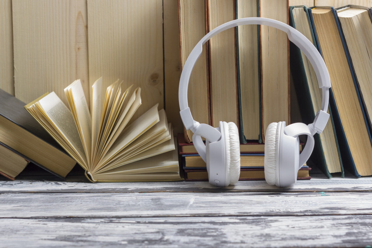 Białe słuchawki nauszne leżące na drewnianym stole przed półką z pionowo ustawionymi książkami. Zdjęcie ilustrujące artykuł pt. audiobooki za darmo na platformie BookBeat