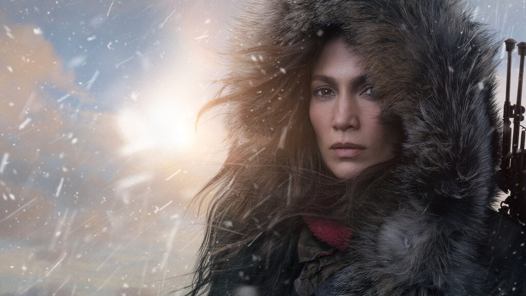 Jenifer Lopez ubrana w grubą, zimową kurtkę z przewieszoną przez ramię bronią. W tle widać padający śnieg i promienie słońca.