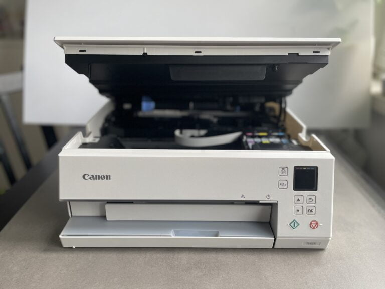 Biała drukarka atramentowa marki Canon z otwartą klapą górną, odsłaniającą wnętrze i pojemniki z tuszem.