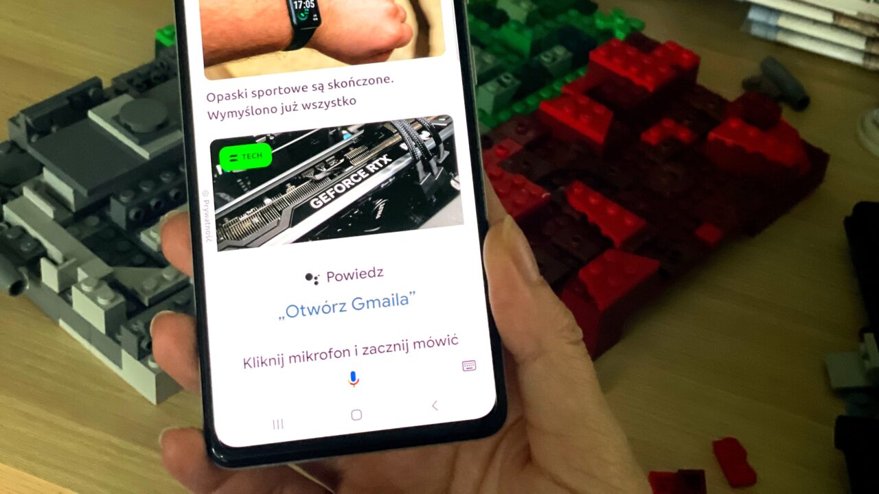 Dłoń trzymająca smartfon z otwartą aplikacją asystenta głosowego, w tle rozmyte klocki LEGO i zegarek na nadgarstku.