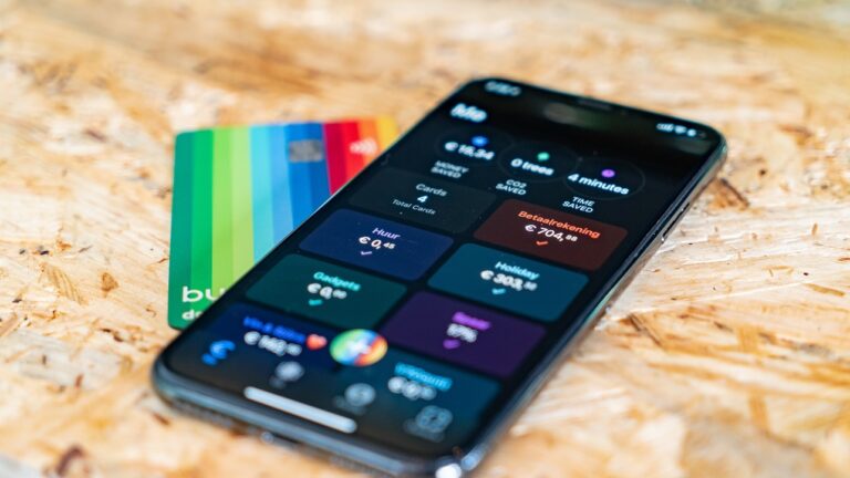 Smartfon z otwartą aplikacją bankową pokazującą informacje o różnych rodzajach transakcji i karty kredytowe w tle na drewnianym blacie.